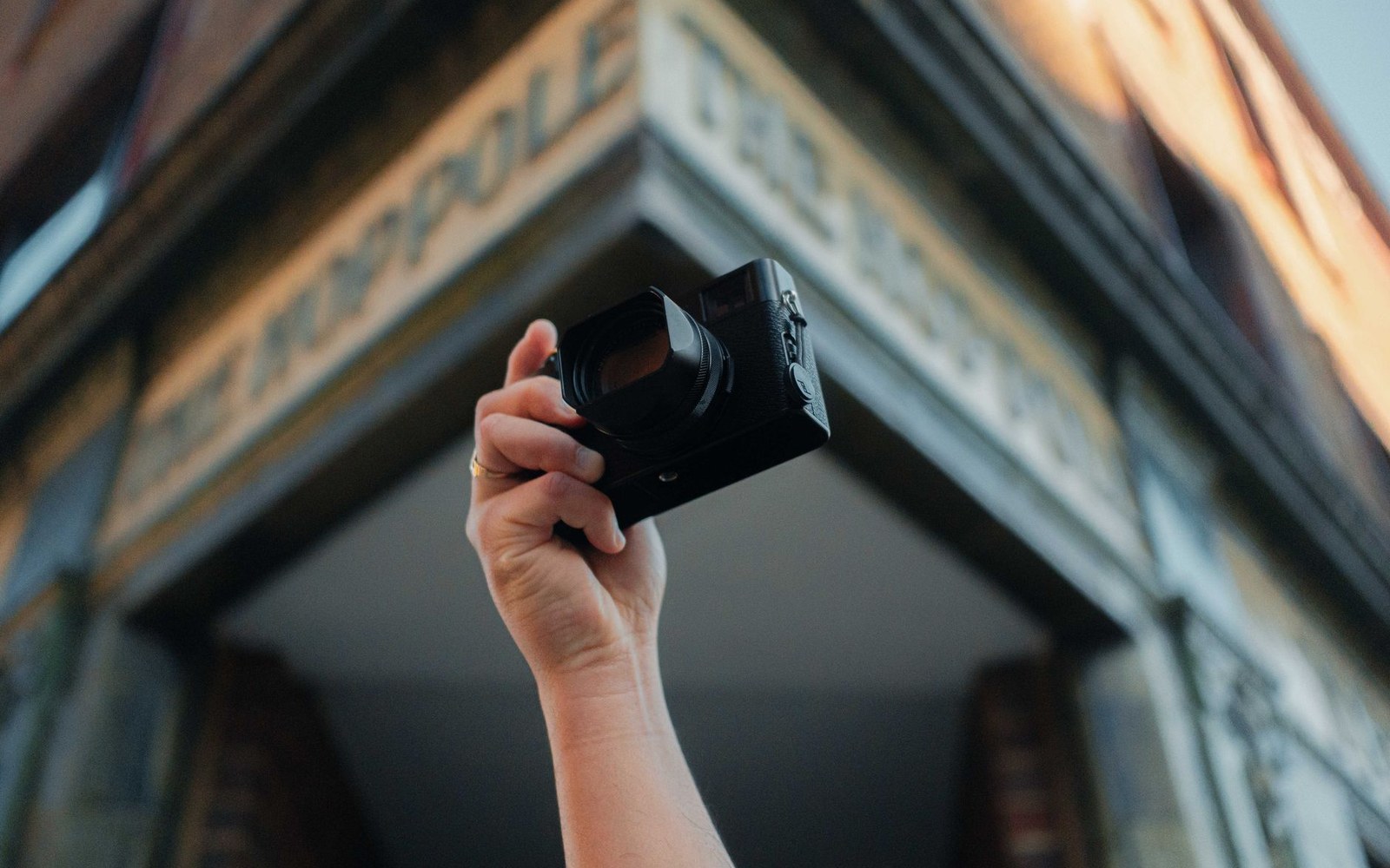 A raised hand holds a compact camera against a building's carved cornice lettering
