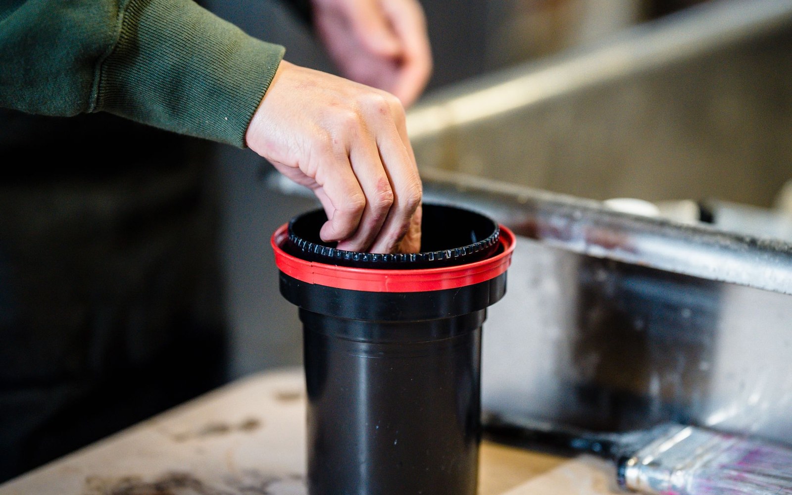 A hand reaches into a black film developing tank with a red lid