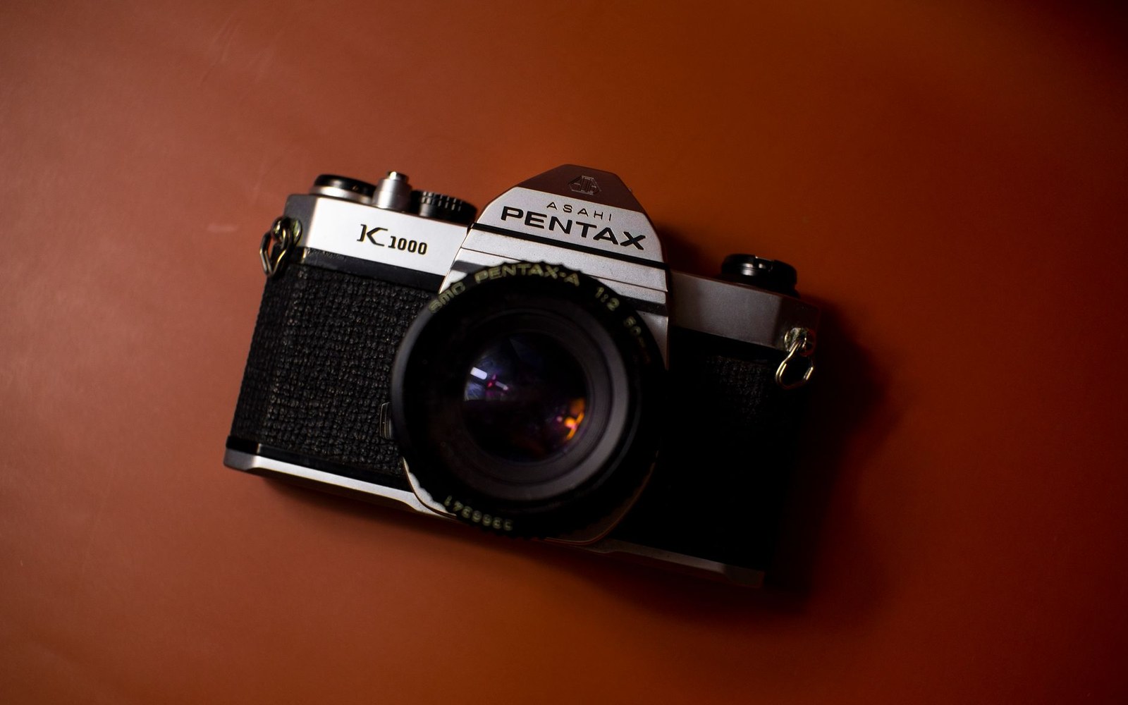 An Asahi Pentax K1000 film camera with lens attached, resting on a reddish-brown surface