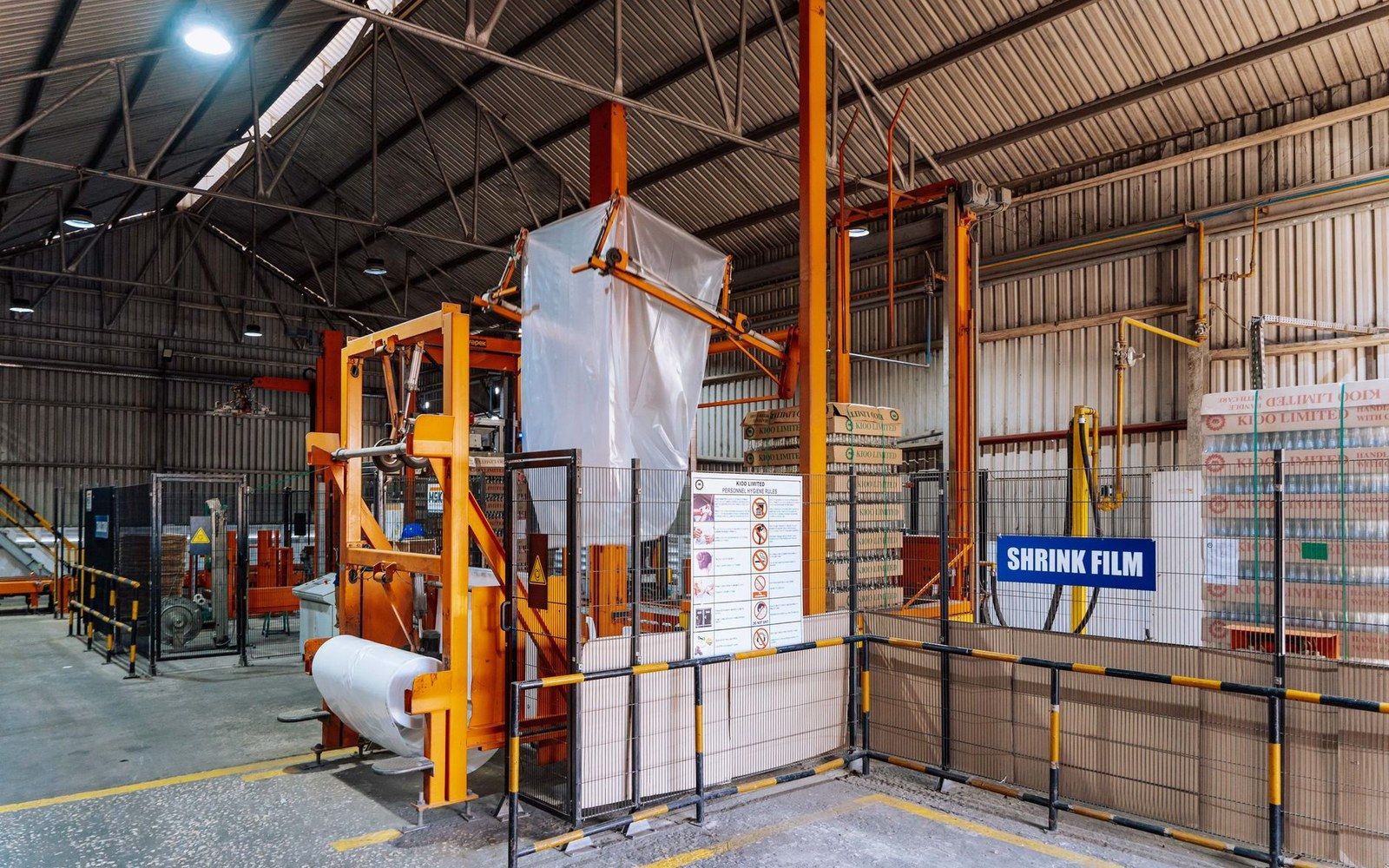 Industrial shrink film wrapping machine surrounded by safety fencing in a warehouse